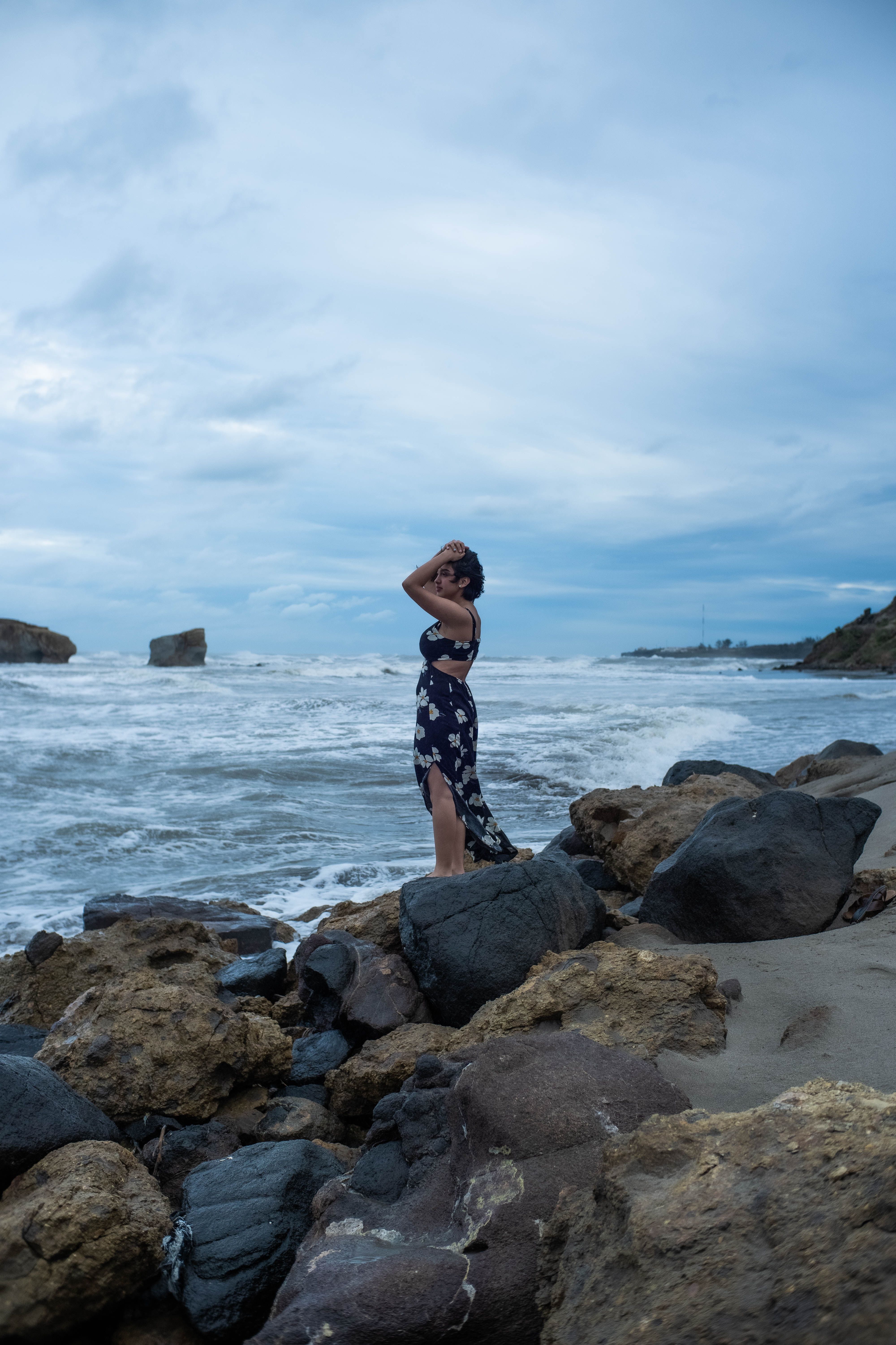 Mulher de vestido longo em pé sobre rochas com ondas quebrando